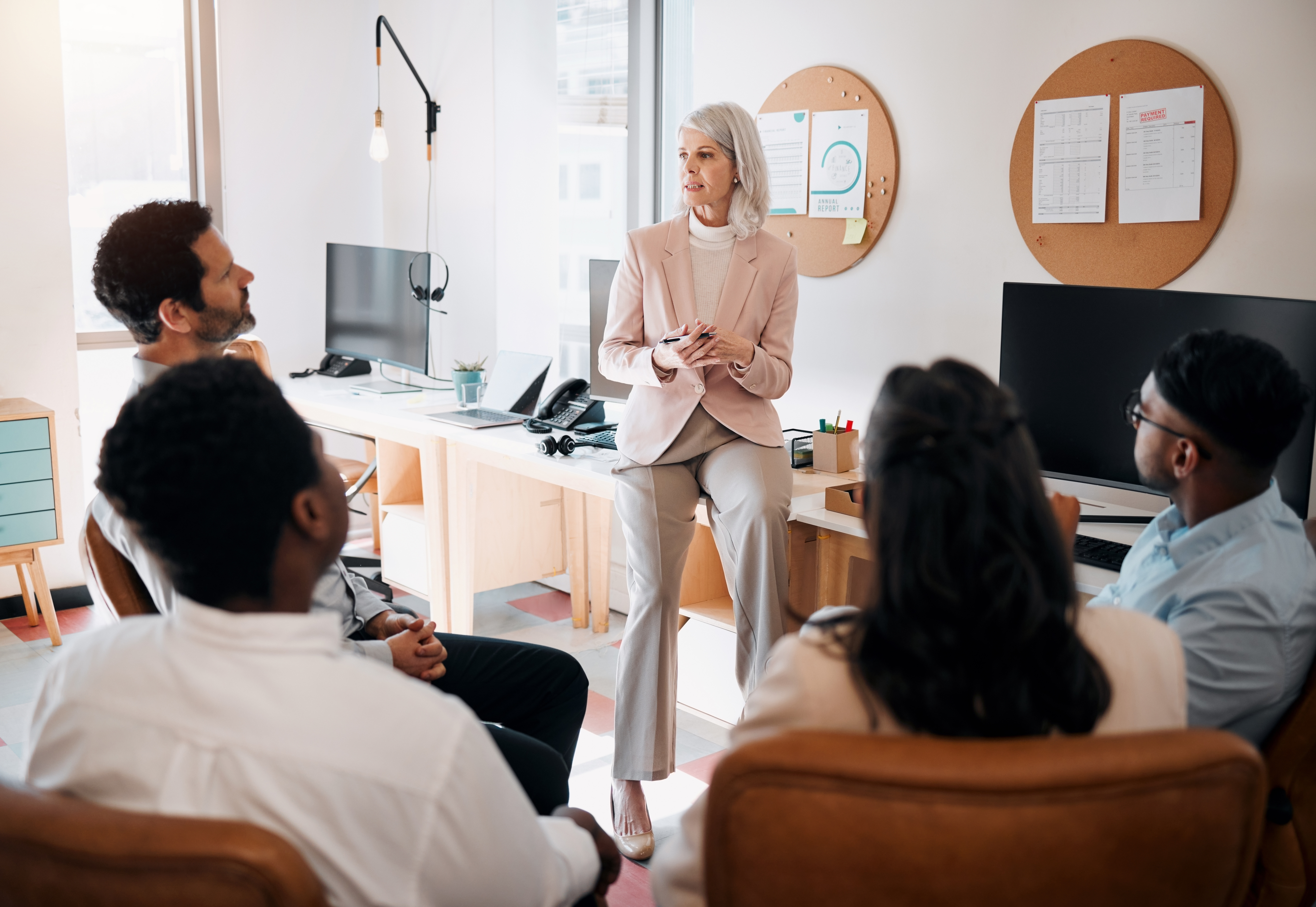 Older female coach sitting on the edge of a long desk talking with four others who are sitting in comfortable chairs.